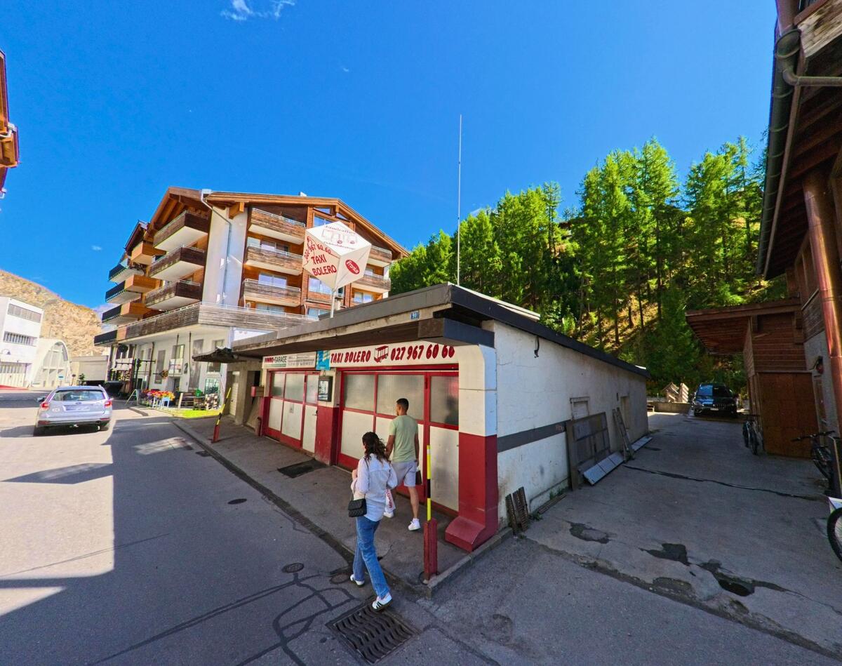 Eine kleine Taxi-Station mit rot-weisser Fassade befindet sich in Zermatt (3920, Kanton Wallis), daneben sind moderne Wohnhaeuser und Waldhaenge zu sehen.