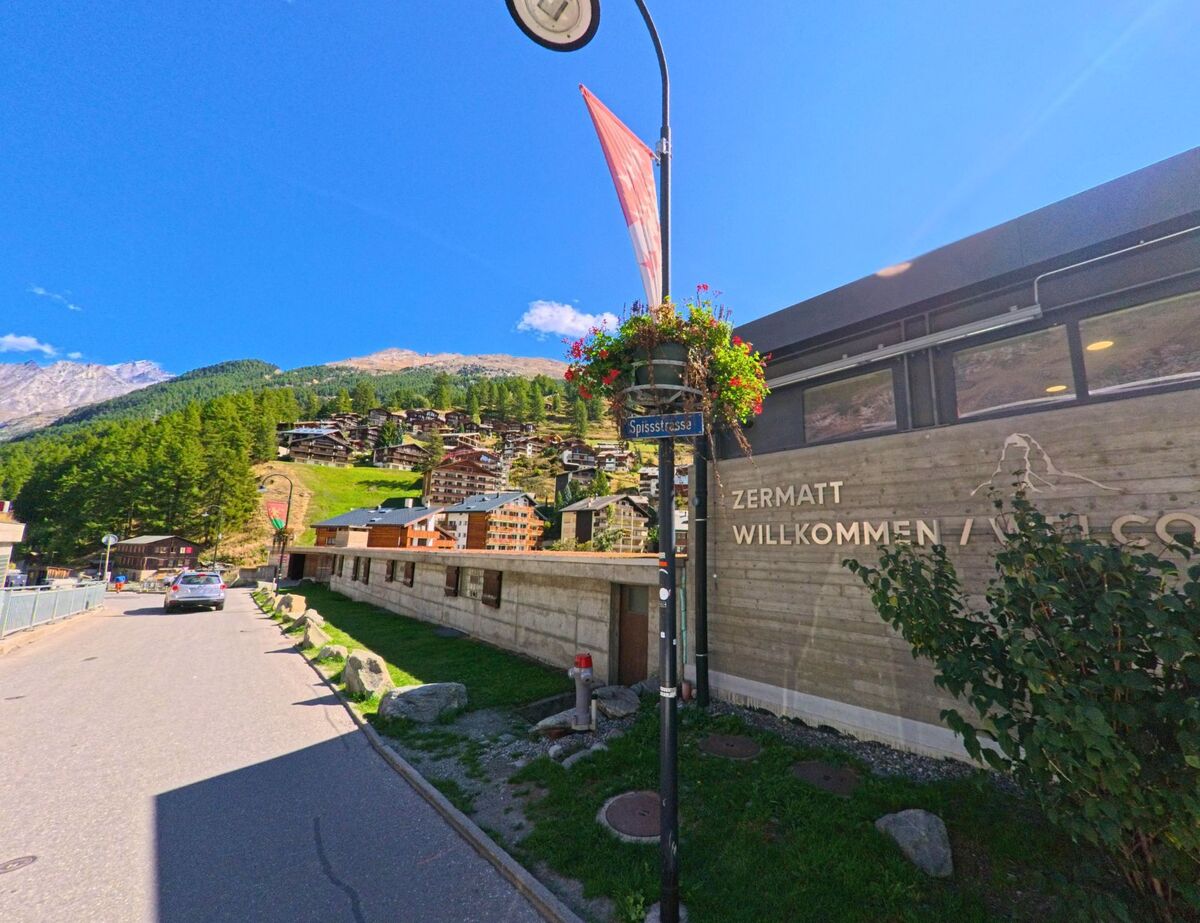 Ein Willkommensschild mit der Aufschrift „Zermatt Willkommen“ steht in Zermatt (3920, Kanton Wallis) an der Spissstrasse, im Hintergrund sind Berghaenge und Chalets sichtbar.