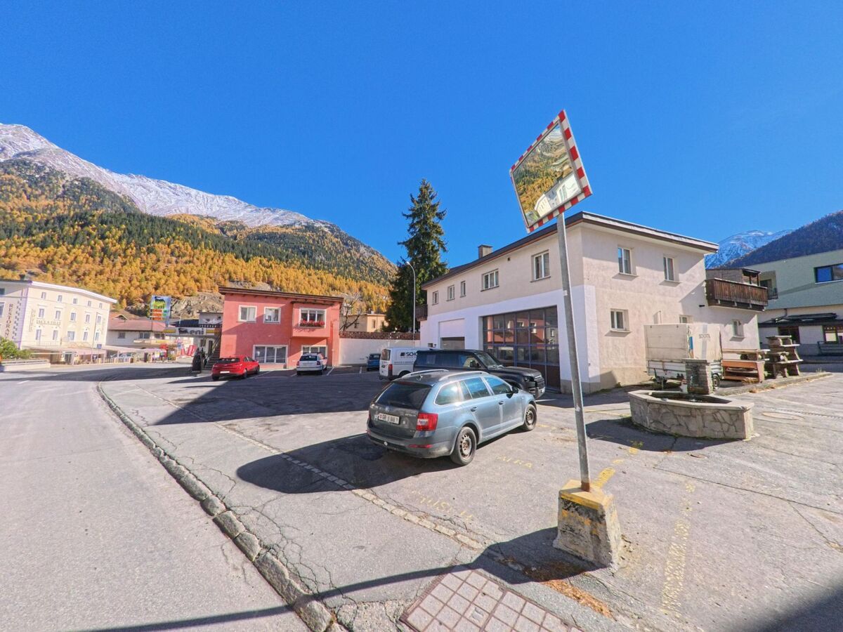 Ein offener Parkplatz mit Wohn- und Gewerbebauten liegt am Rand des Dorfzentrums von Zernez (7530, Kanton Graubünden) vor herbstlich gefärbten Berghängen unter klarem Himmel.