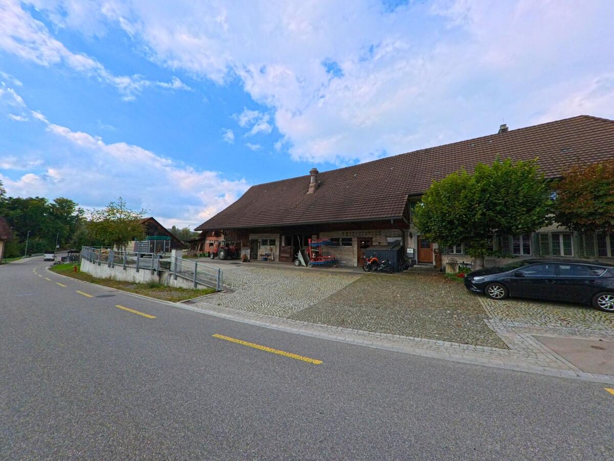 In Zetzwil (5732, Kanton Aargau) liegt ein grosser Bauernhof mit Pflasterplatz und landwirtschaftlichen Geraeten an einer sanft geschwungenen Strasse.