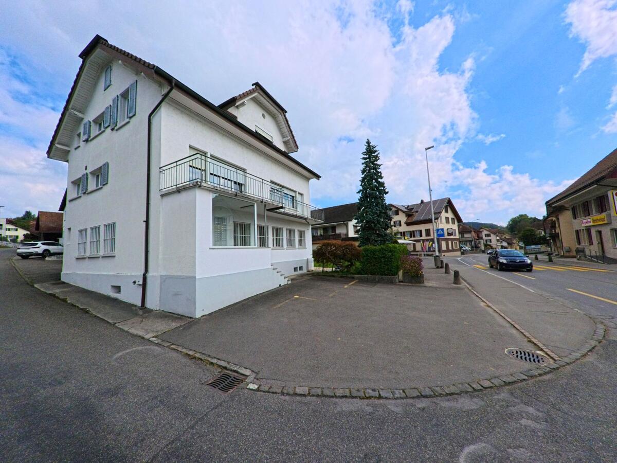 In Zetzwil (5732, Kanton Aargau) steht ein weisses Haus mit Balkon an einer Dorfstrasse, die von weiteren Wohnhaeusern und einem Nadelbaum gesaeumt wird.