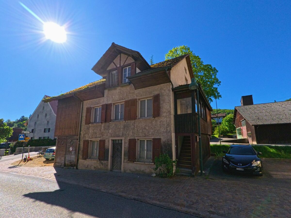 Ein älteres Wohnhaus mit Holzverkleidung und roten Fensterläden steht an einer Kreuzung in Ziefen (4417, Kanton Basel-Landschaft), beleuchtet von der tief stehenden Sonne.