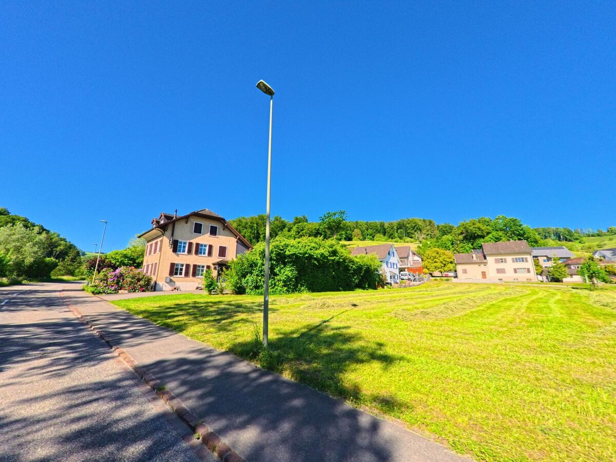 In Ziefen (4417, Kanton Basel-Landschaft) steht ein freistehendes Haus mit Fensterläden am Rand frisch gemähter Wiesen, umgeben von weiteren Gebäuden und Wald.