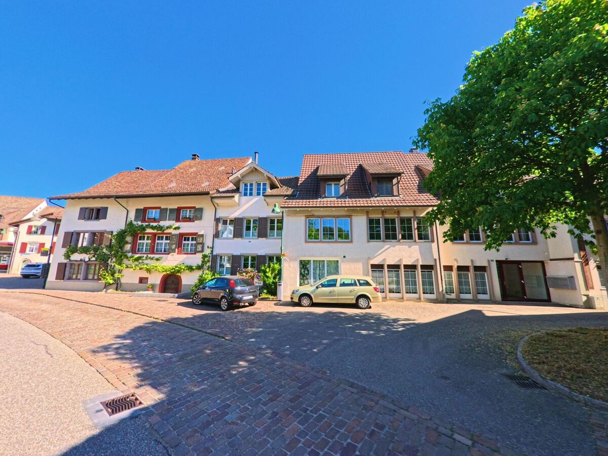 Am Rand eines Platzes in Ziefen (4417, Kanton Basel-Landschaft) stehen gepflegte Wohnhäuser mit begrünten Fassaden und davor parkenden Autos.