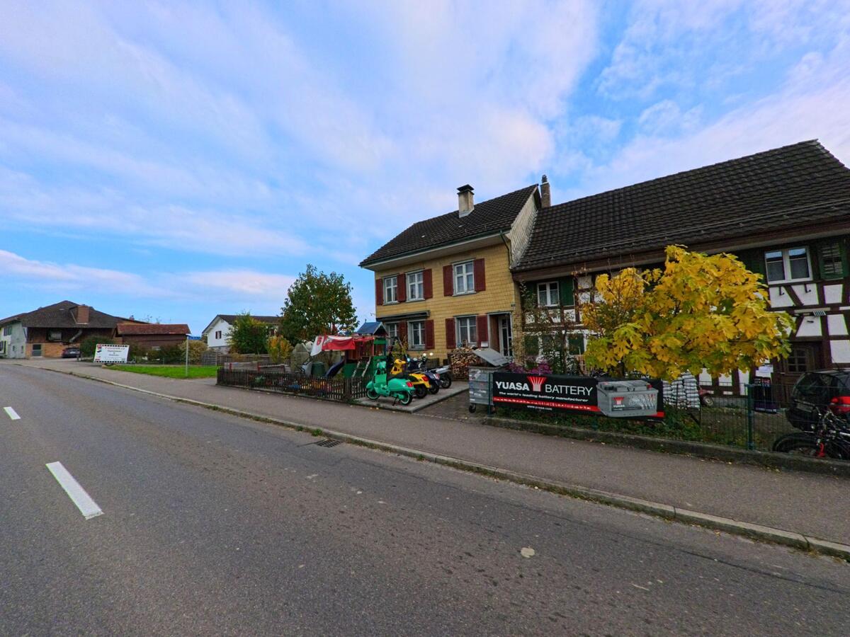 An einer Dorfstrasse in Zihlschlacht-Sitterdorf (8588, Kanton Thurgau) stehen ein Riegelhaus und ein Wohnhaus mit kleinen Vorgärten, davor parkierte Motorroller.