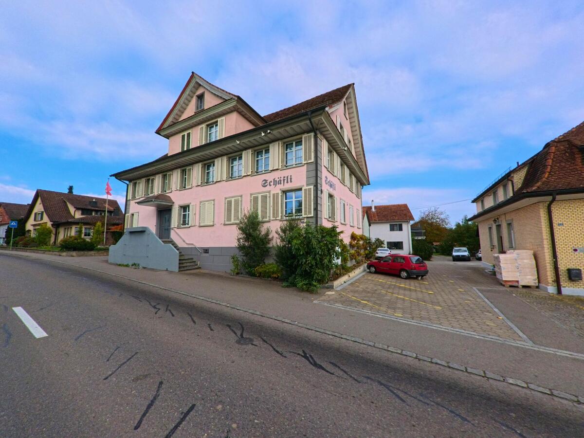 An einer Strasse in Zihlschlacht-Sitterdorf (8588, Kanton Thurgau) steht ein historisches Gasthaus mit der Aufschrift Schäfli, umgeben von Wohnhäusern und Parkplätzen.