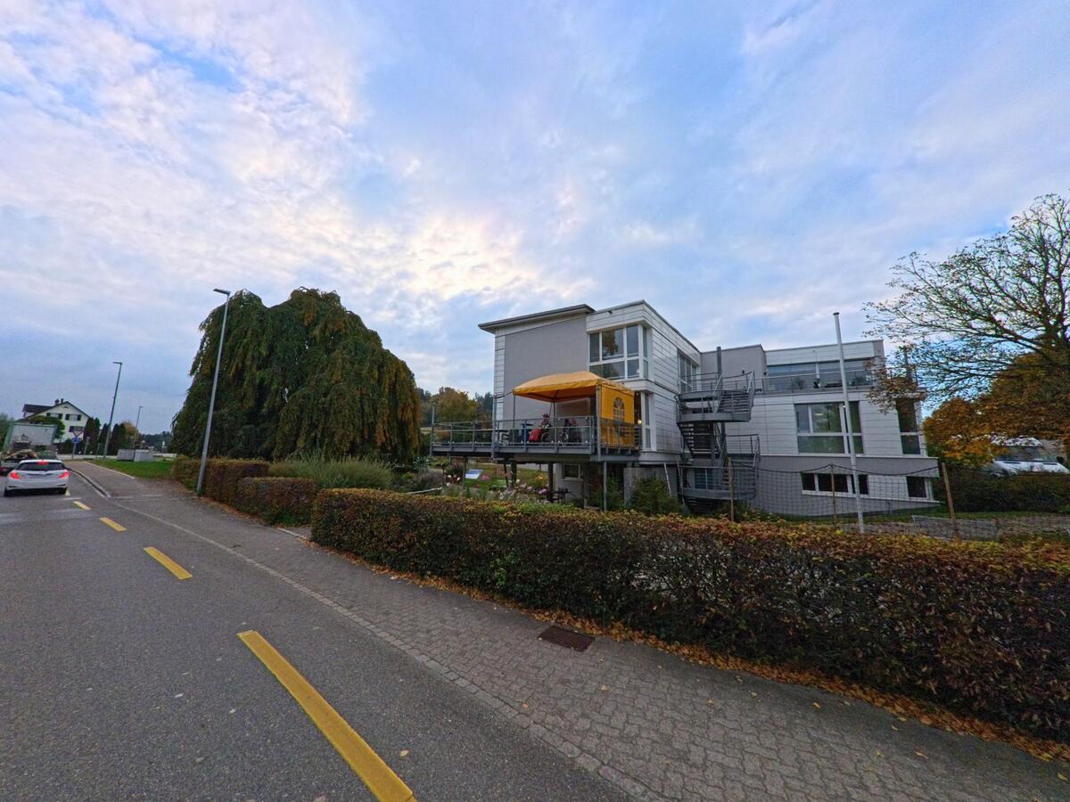 Neben einem Gehweg in Zihlschlacht-Sitterdorf (8588, Kanton Thurgau) steht ein modernes Gebäude mit Terrasse und Treppenanlage, umgeben von Hecken und herbstlicher Bepflanzung.