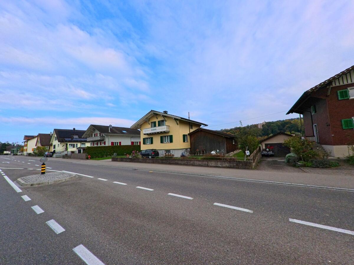 An einer breiten Strasse in Zihlschlacht-Sitterdorf (8588, Kanton Thurgau) reihen sich Wohnhäuser mit Gärten und Garagen entlang eines leicht ansteigenden Quartiers.