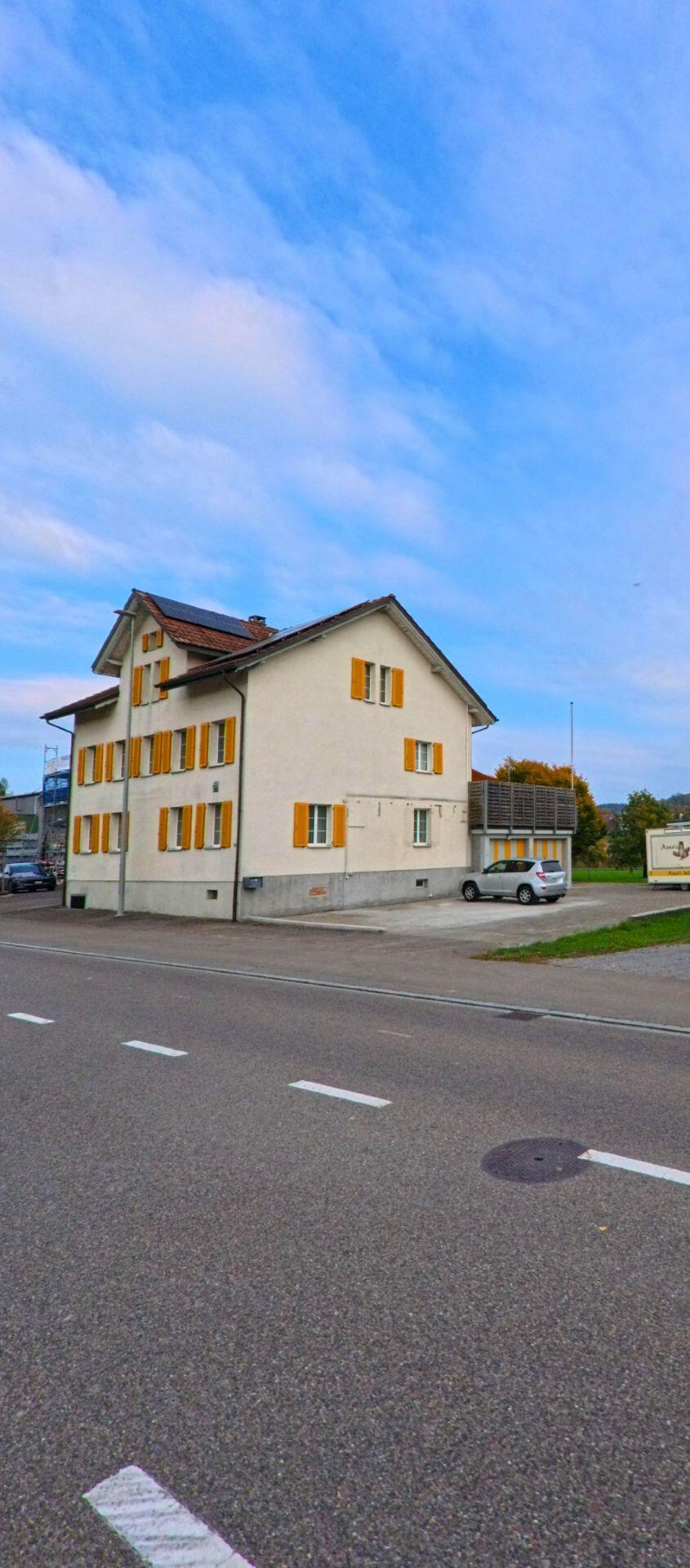 An einer Hauptstrasse in Zihlschlacht-Sitterdorf (8588, Kanton Thurgau) steht ein helles Wohnhaus mit gelben Fensterläden am Rand einer offenen Umgebung.