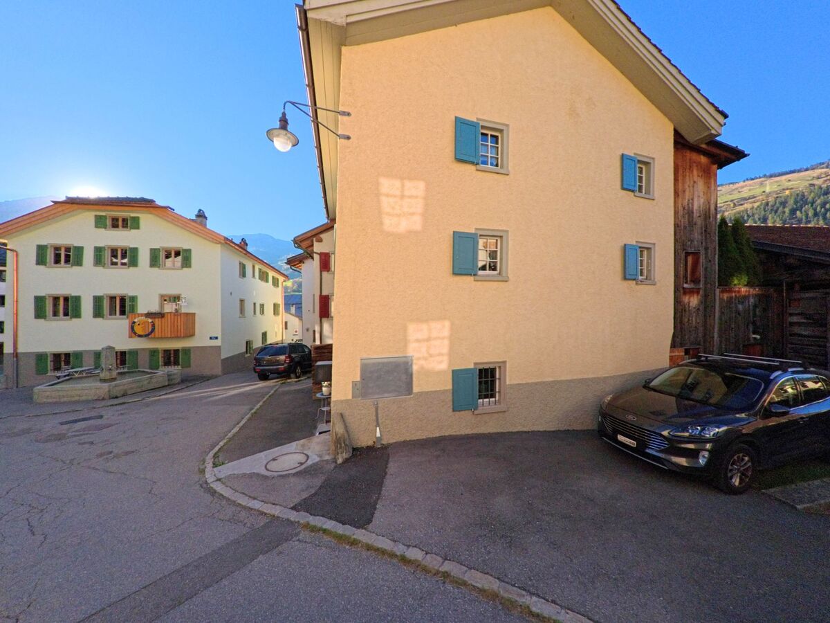 An einer kleinen Dorfecke mit schmaler Fahrbahn stehen Wohnhäuser mit farbigen Fensterläden in Zillis-Reischen (7432, Kanton Graubünden), während eine Strassenlampe die Kreuzung markiert.