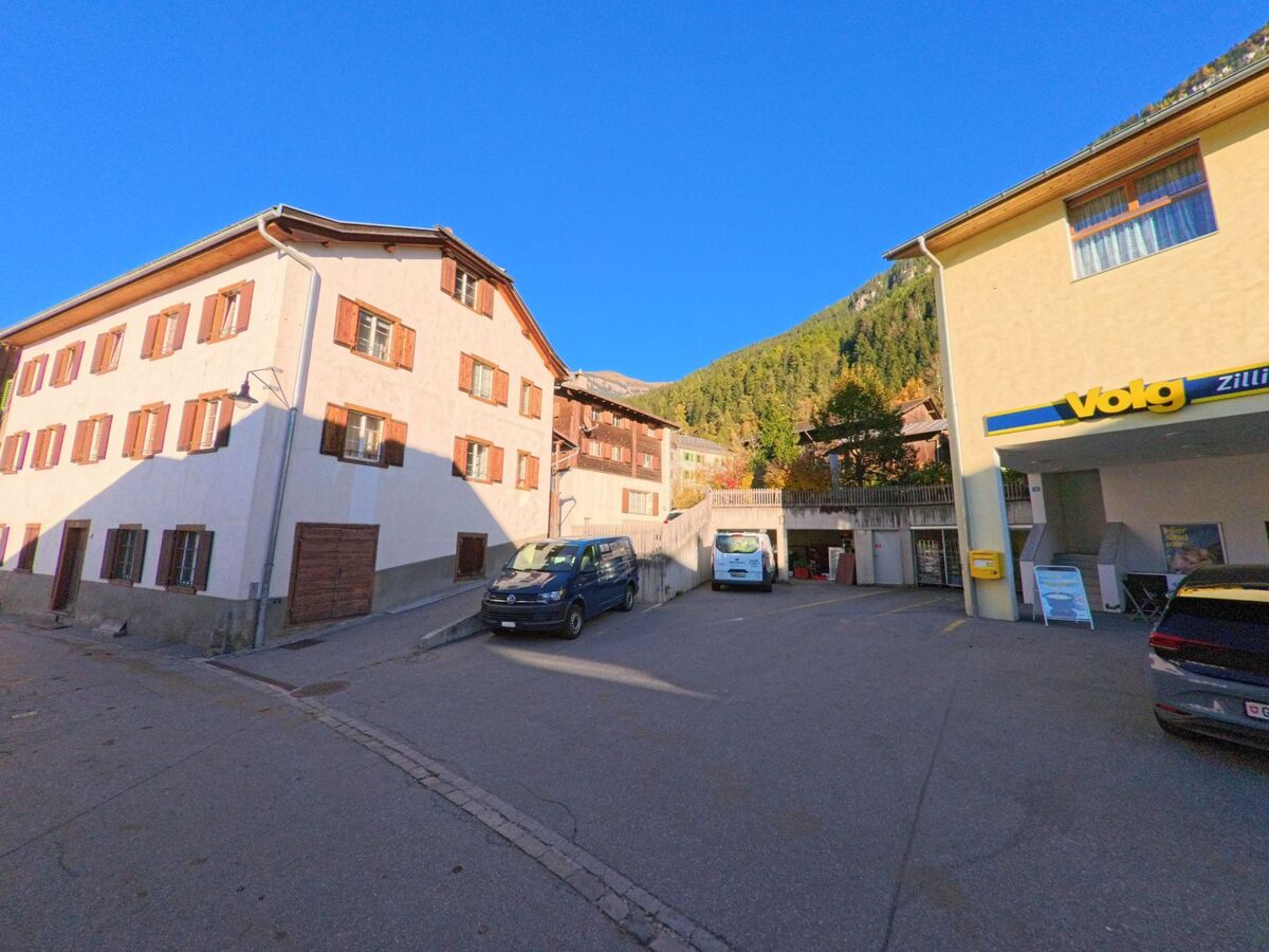 Vor einem kleinen Dorfplatz mit Wohnhäusern und parkenden Fahrzeugen befindet sich in Zillis-Reischen (7432, Kanton Graubünden) ein lokales Geschäft vor bewaldeten Berghängen.