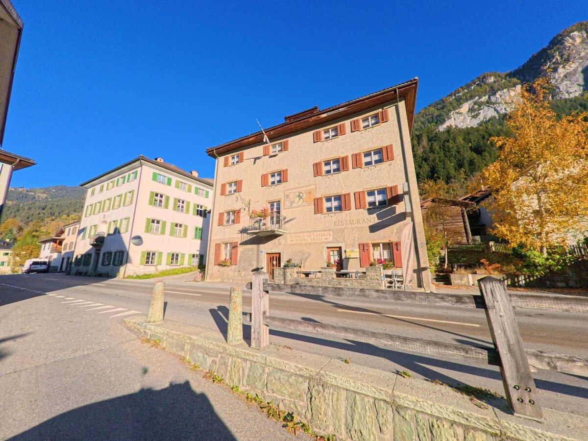 An einer Durchgangsstrasse mit historischen Wohn- und Gasthausfassaden zeigt sich das Dorfzentrum von Zillis-Reischen (7432, Kanton Graubünden) vor steil ansteigenden Berghängen.