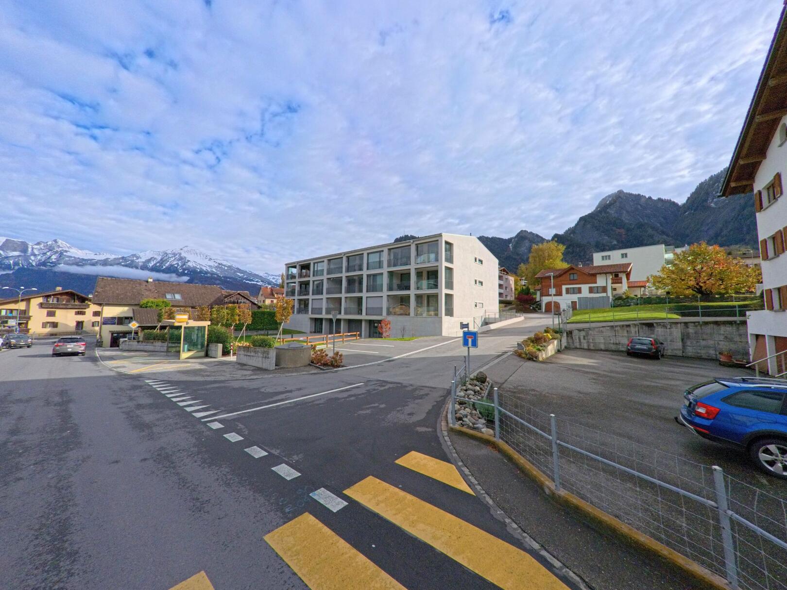An einer übersichtlichen Strassenkreuzung in Zizers (7205, Kanton Graubünden) steht ein modernes Wohngebäude, eingerahmt von Einfamilienhäusern und einer alpinen Bergkulisse im Hintergrund.