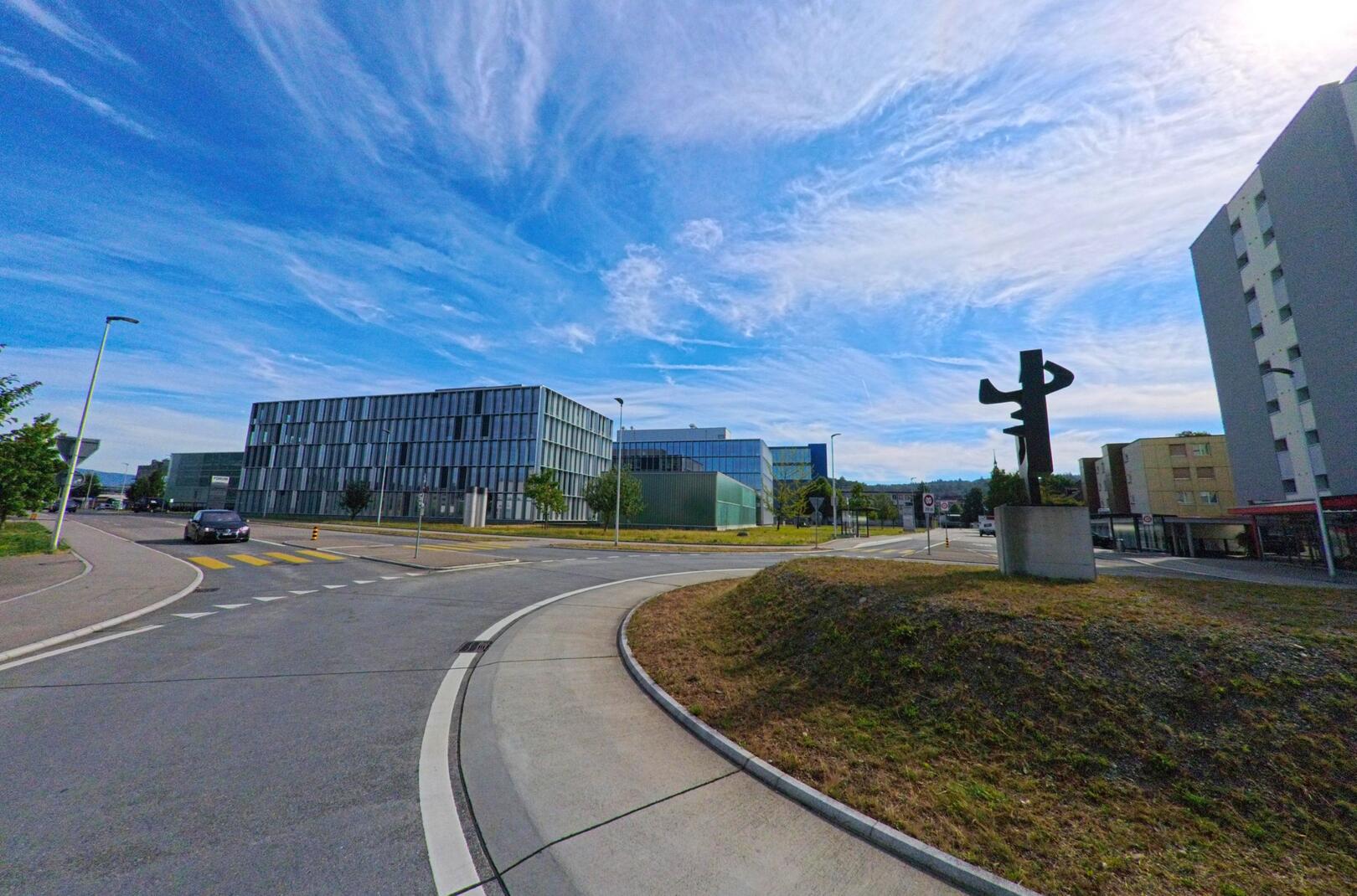 In Zofingen (4800, Kanton Aargau) steht eine moderne Skulptur auf einem Verkehrsinselhügel, umgeben von zeitgenoessischer Architektur und Mehrfamilienhaeusern.
