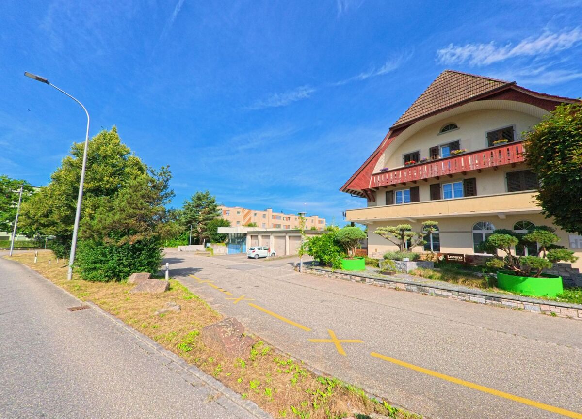 In Zofingen (4800, Kanton Aargau) steht ein traditionelles Wohnhaus mit rotem Balkon vor einer gepflegten Gartenanlage und moderner Wohnbebauung im Hintergrund.