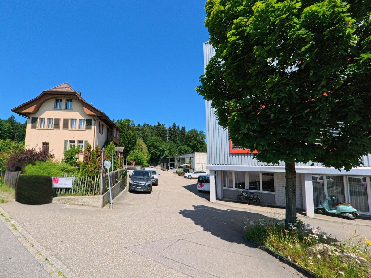 In Zollikofen (3052, Kanton Bern) trifft ein historisches Wohnhaus mit markantem Dach auf moderne Gewerbebauten entlang einer kleinen Nebenstrasse mit geparkten Fahrzeugen und Bäumen.