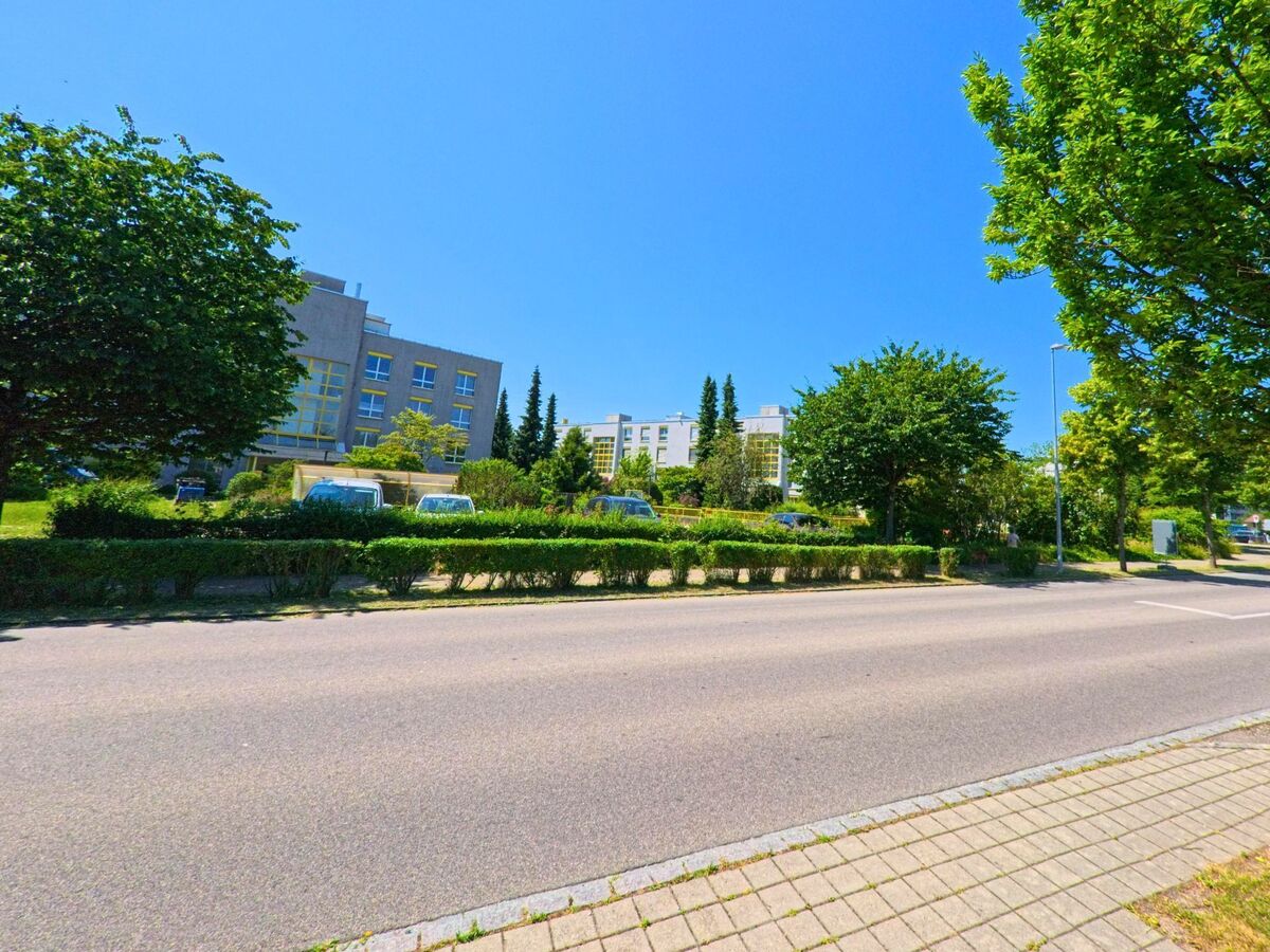 Entlang einer Hauptstrasse in Zollikofen (3052, Kanton Bern) stehen moderne Wohnhäuser hinter einer begrünten Parkzone mit Hecken, Bäumen und parkenden Autos.