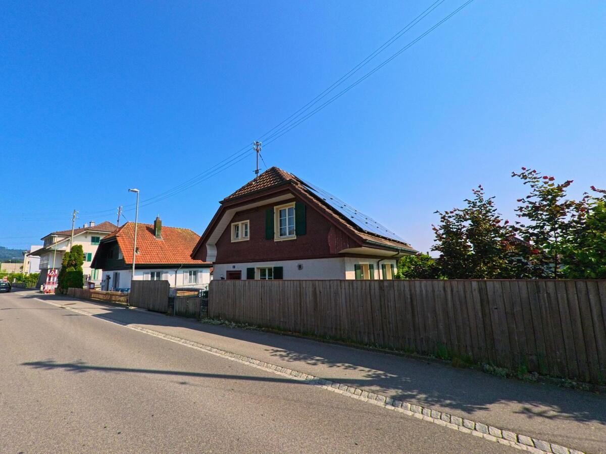 In Zollikofen (3052, Kanton Bern) reihen sich entlang einer ruhigen Quartierstrasse mehrere Einfamilienhäuser mit Ziegeldächern, von denen eines mit Solarpanels ausgestattet ist.