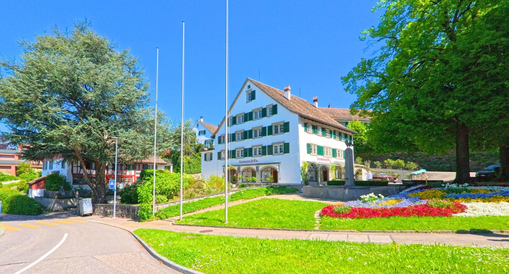 Im Zentrum von Zollikon, PLZ 8702 im Kanton Zürich, steht ein traditionsreiches Gebäude mit grün-weissen Fensterläden neben einem gepflegten Blumenbeet und grossen Bäumen.