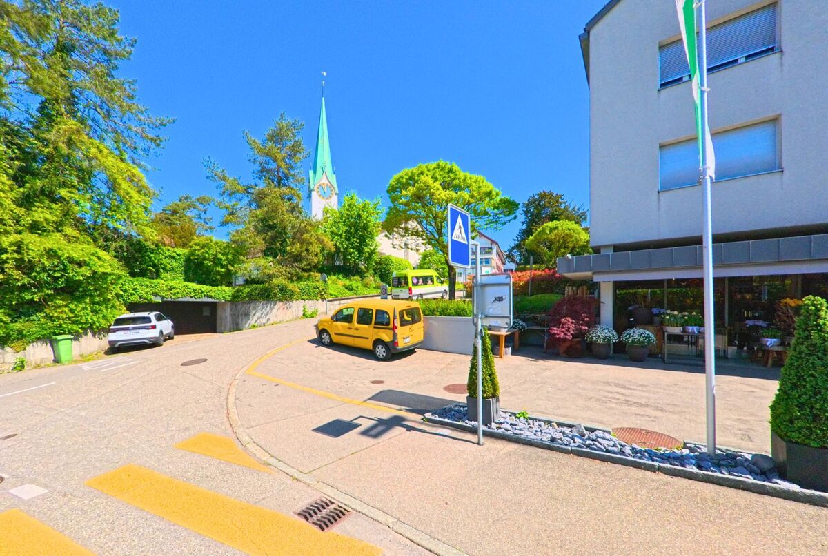 In Zollikon, PLZ 8702 im Kanton Zürich, erhebt sich hinter einem Wohnquartier mit Geschäften der grüne Kirchturm der reformierten Kirche über Bäumen und Dächern.