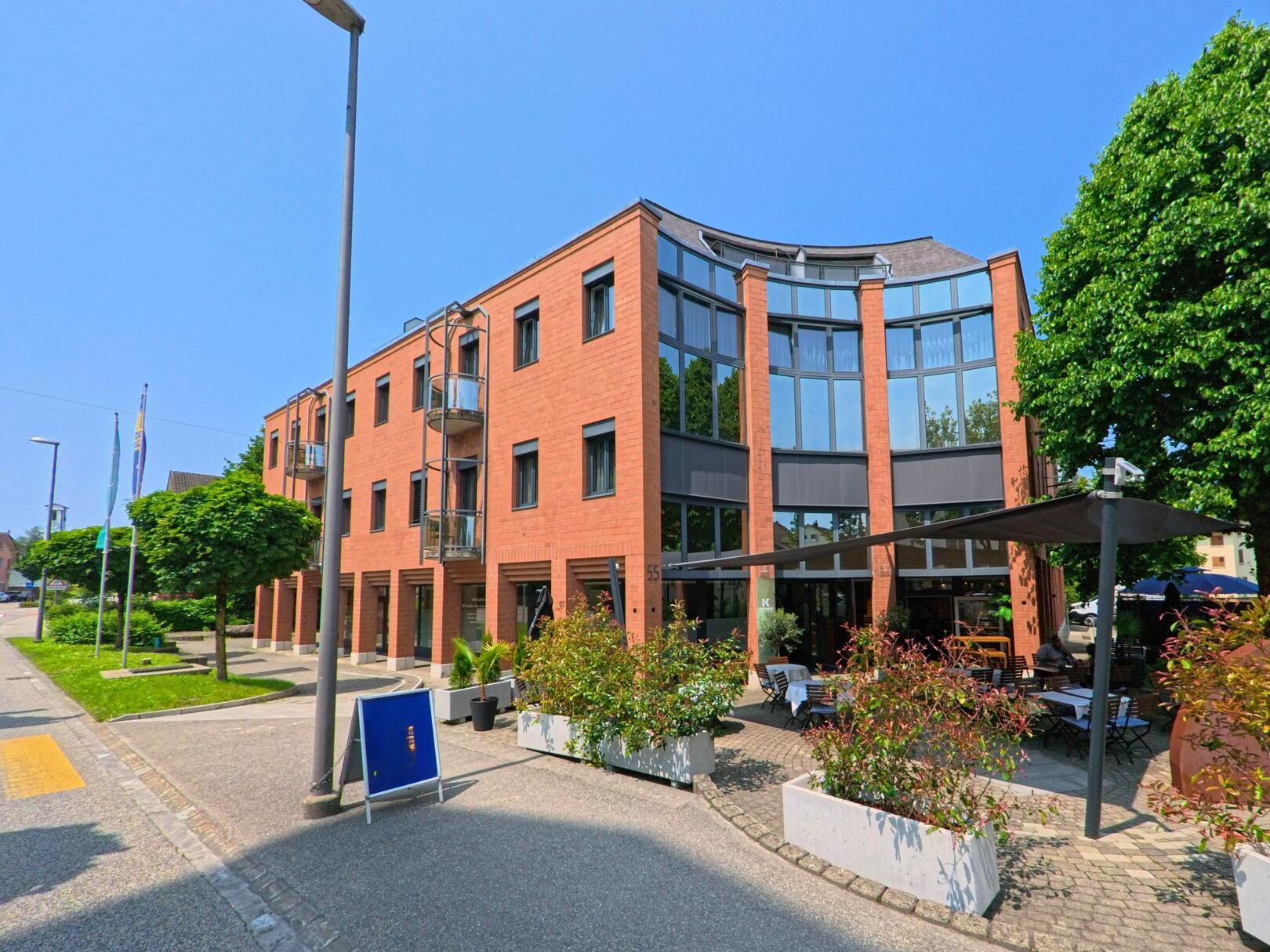 Das moderne Gebäude in Zuchwil (4528, Kanton Solothurn) zeigt eine rot-braune Fassade mit grossen Fenstern und einem Restaurantbereich im Vordergrund, der von Pflanzen umgeben ist.