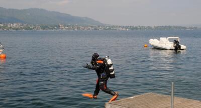 Am Ufer des Sees springt in Küsnacht (8127, Kanton Zürich) eine Person in Tauchausrüstung von einem Steg ins Wasser, waehrend Boote auf dem ruhigen See treiben und das andere Ufer sichtbar ist.
