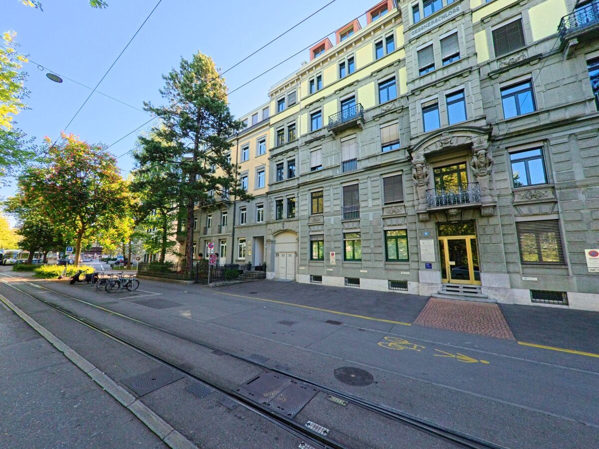 In Zürich 8021 im Kanton Zürich steht ein historisches Gebäude mit aufwendig verzierter Fassade direkt an einer Strasse mit Tramgleisen und Baumbestand.