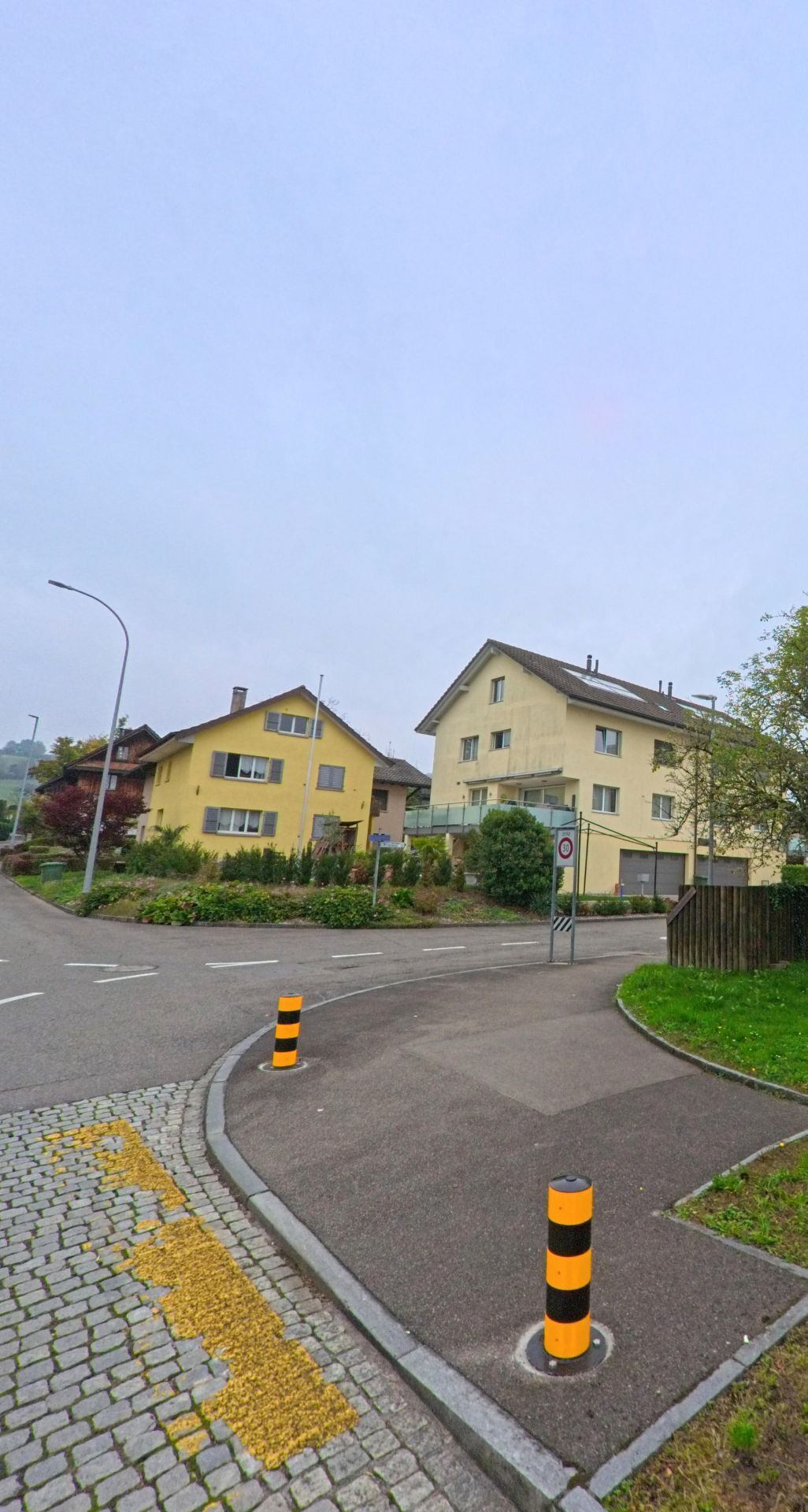 In Zufikon (5621, Kanton Aargau) stehen zwei hellgelbe Wohnhaeuser an einer leicht gekruemmten Strasse mit markierten Pollern und gepflegten Beeten.