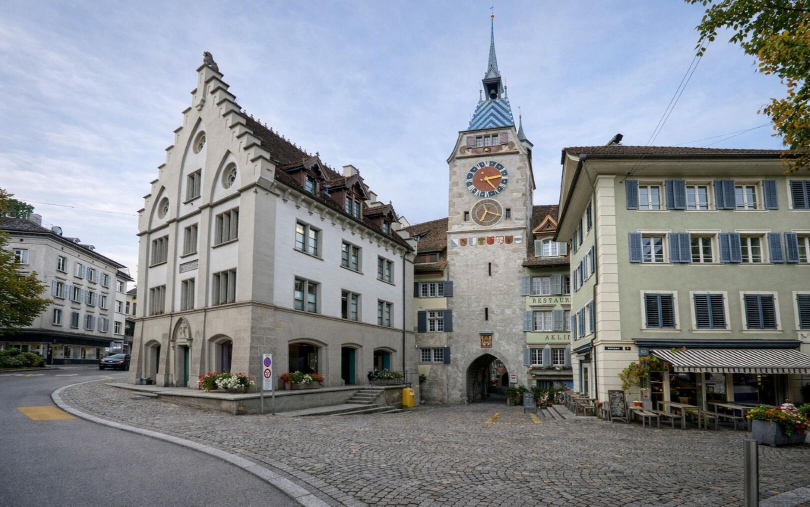 In der Altstadt von Zug (6300, Kanton Zug) praegt ein historischer Turm mit farbiger Uhr das gepflasterte Platzbild zwischen eng stehenden traditionellen Gebaeuden.