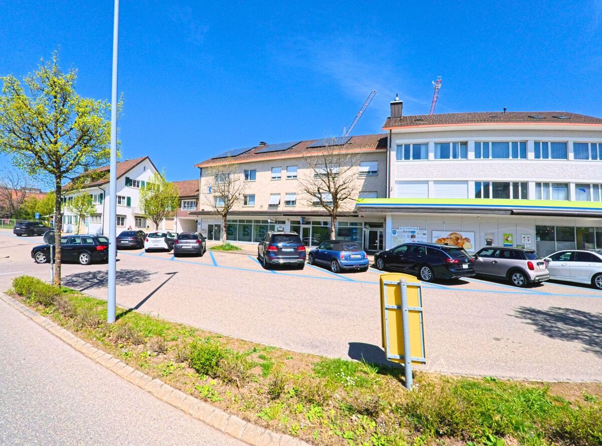 Vor einem kleinen Einkaufszentrum in Zumikon, PLZ 8126 im Kanton Zürich, stehen mehrere Autos auf markierten Parkplätzen an einem sonnigen Tag.