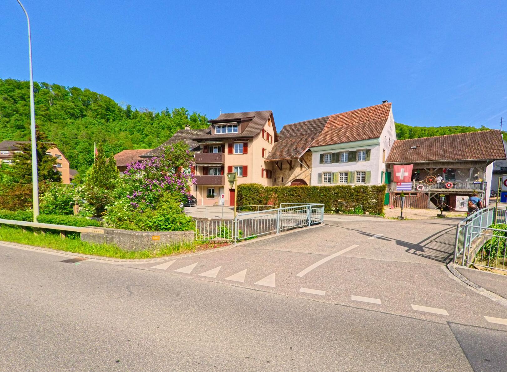 In Zunzgen (4455, Kanton Baselland) fuehrt eine Strasse ueber einen kleinen Bachlauf hin zu historischen Wohnhaeusern mit markanten Fassaden am Dorfrand.