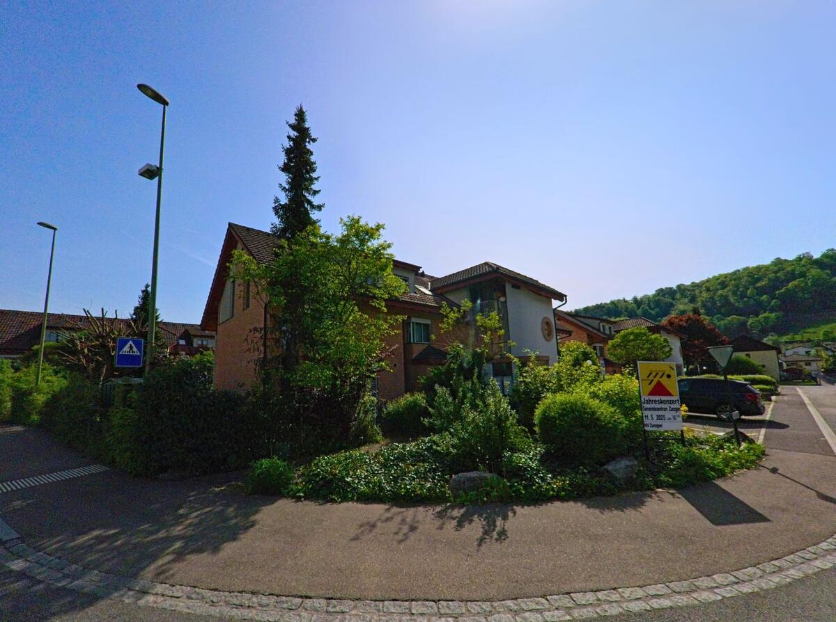In Zunzgen (4455, Kanton Baselland) zweigt eine Wohnstrasse mit Einfamilienhaeusern und gepflegten Gaerten an einem Veranstaltungsplakat in Richtung Siedlung ab.