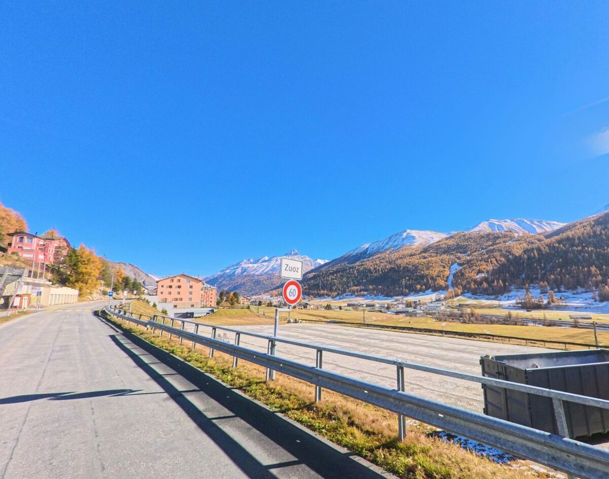 Am Ortseingang von Zuoz (7524, Kanton Graubünden) verläuft eine Strasse entlang offener Flächen, wobei das Ortsschild und die alpine Berglandschaft im Hintergrund sichtbar sind.