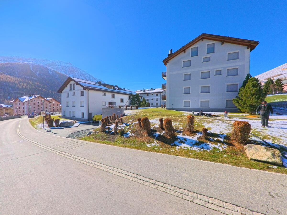 Entlang einer leicht ansteigenden Strasse in Zuoz (7524, Kanton Graubünden) stehen mehrere Wohnhäuser mit gepflegten Grünflächen und vereinzelten Schneeresten vor der alpinen Bergkulisse.