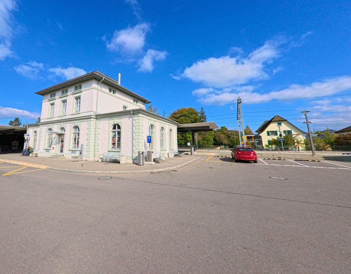 Am Bahnhof von Zurzach (5330, Kanton Aargau) steht ein helles historisches Gebaeude neben Gleisanlagen und einem kleinen Vorplatz mit parkenden Autos.