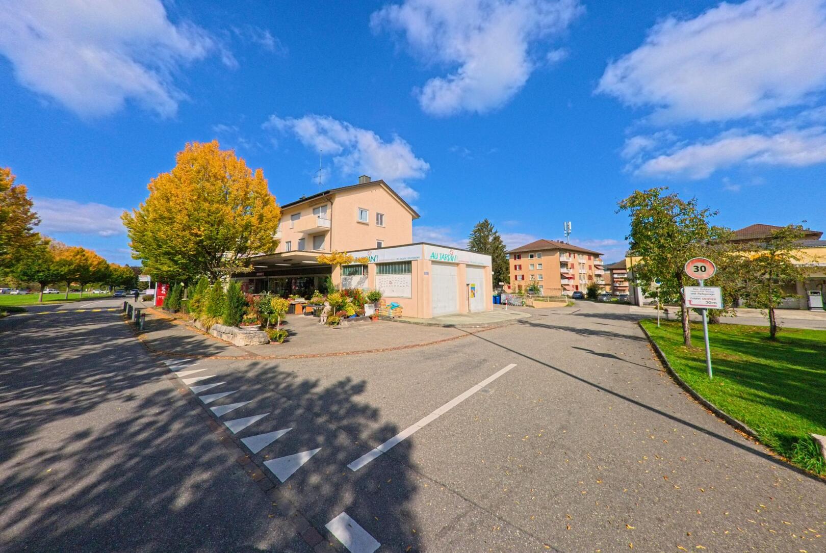 In Zurzach (5330, Kanton Aargau) praegt eine kleine Ladenzeile mit Wohnhaeusern und herbstlichen Baumen das ruhige Strassenbild einer Tempo-30-Zone.
