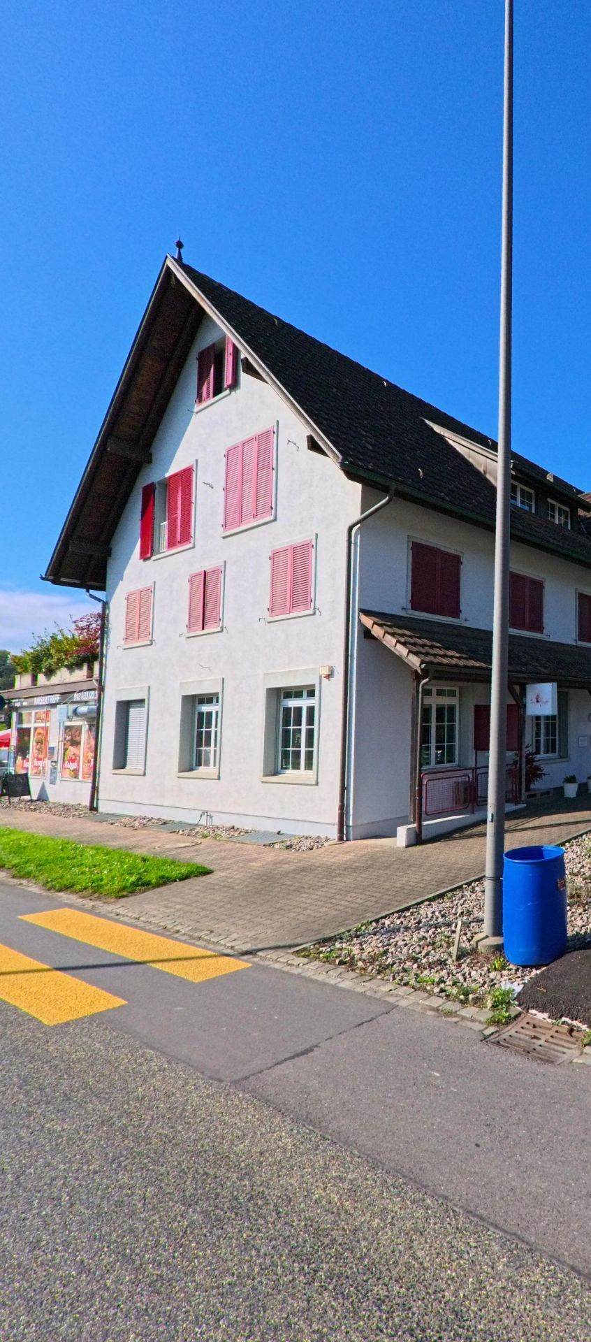 In Zurzach (5466, Kanton Aargau) steht an einer Strasse mit Zebrastreifen ein Haus mit roten Fensterlaeden und kleinem Ladeneingang.