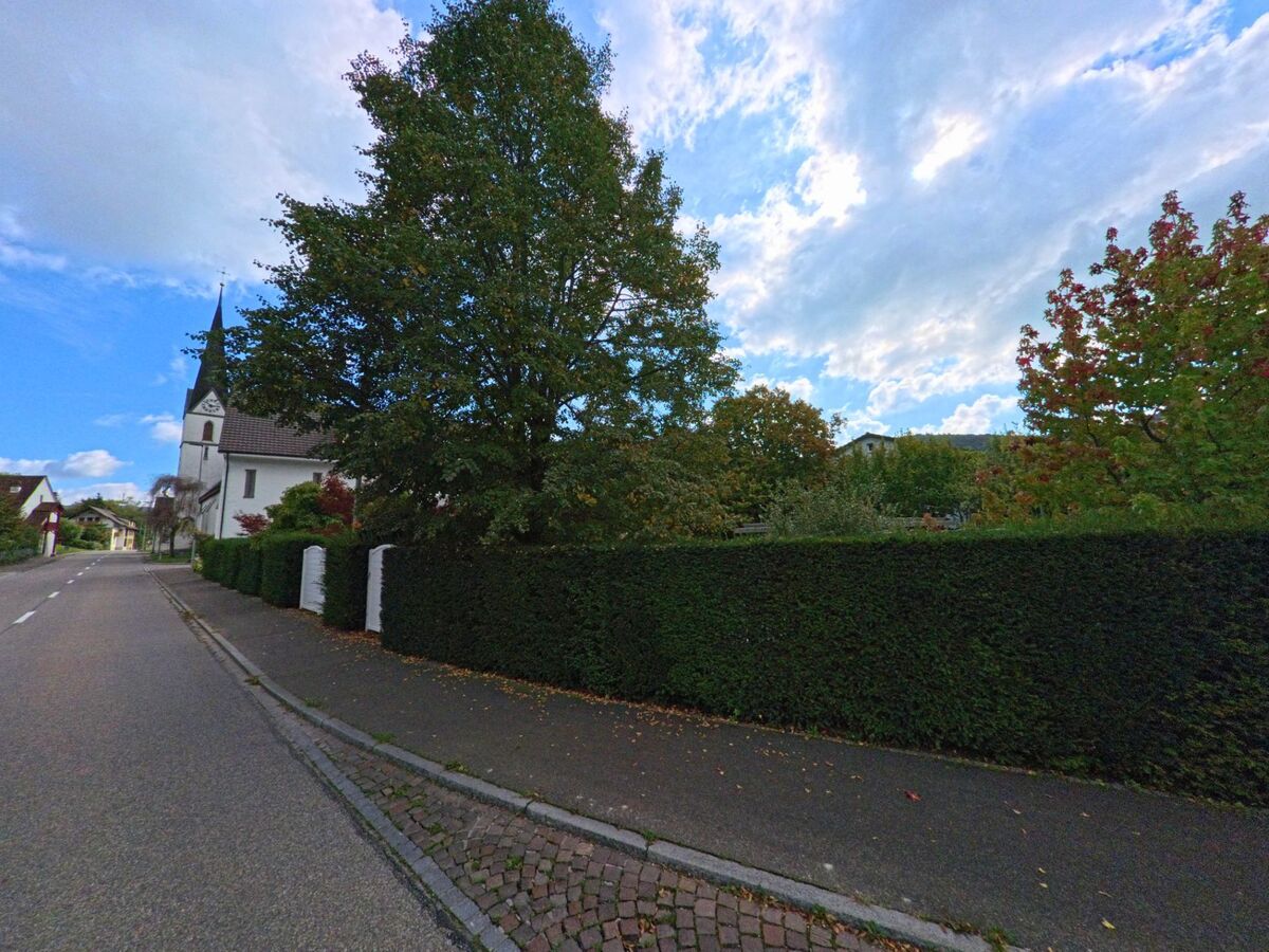 In Zuzgen (4315, Kanton Aargau) fuehrt eine ruhige Strasse an einer Kirche mit schlankem Turm vorbei, flankiert von gepflegten Hecken und Baeumen.