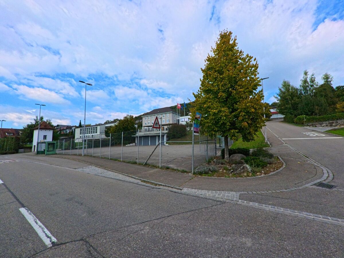 In Zuzgen (4315, Kanton Aargau) liegt ein grosser geteerrter Platz mit Zaun und Schulgebaeuden neben einer leicht ansteigenden Strasse und einem einzeln stehenden Baum.