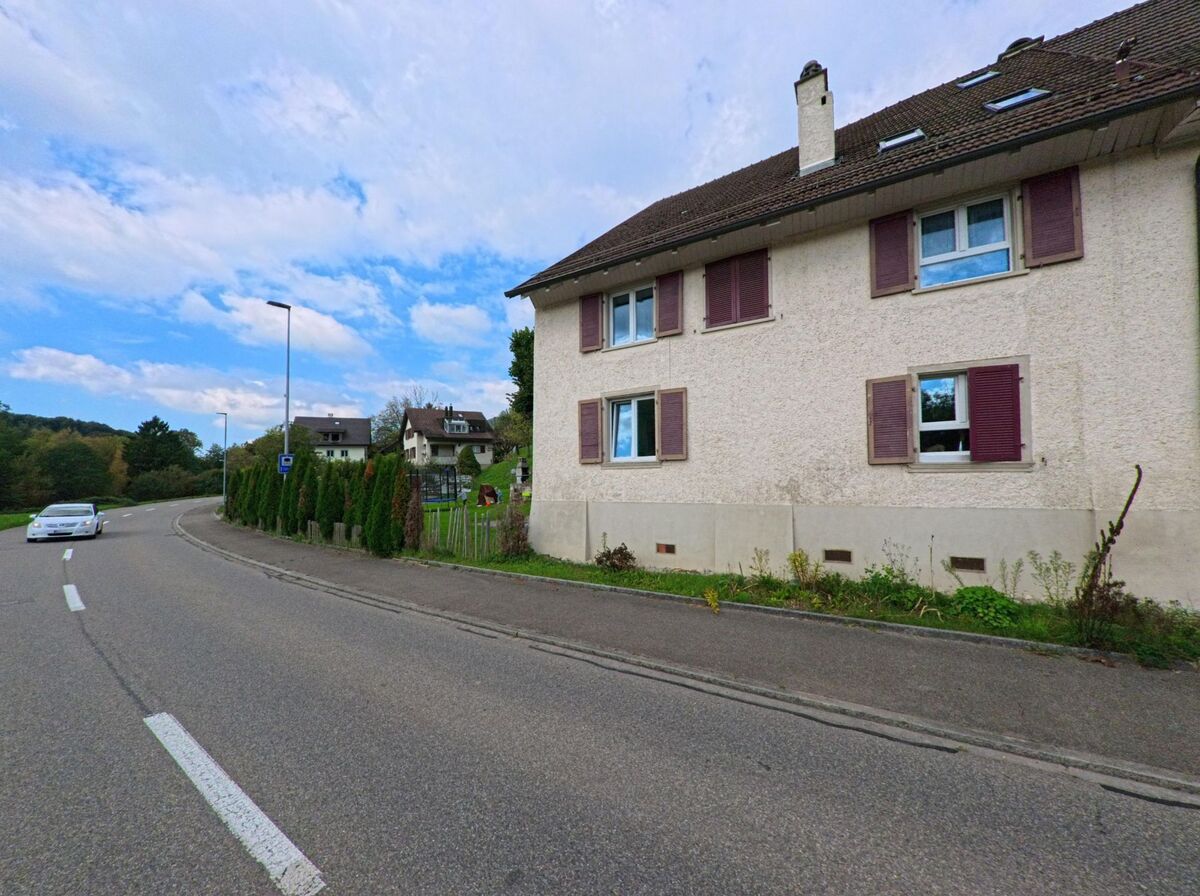 In Zuzgen (4315, Kanton Aargau) verläuft eine sanft gekruemmte Strasse an einem Haus mit violetten Fensterlaeden vorbei, umgeben von vereinzelten Baeumen und weiteren Wohnhaeusern.