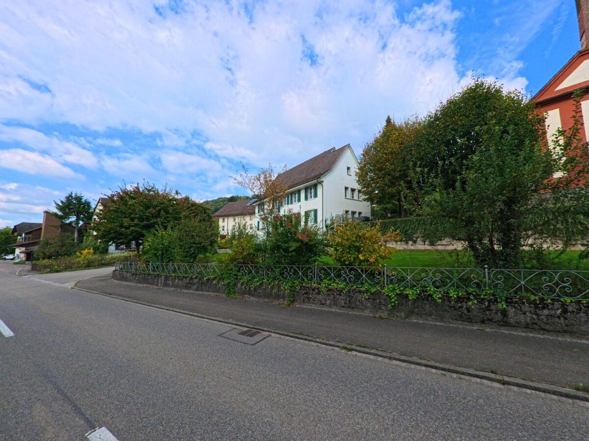 In Zuzgen (4315, Kanton Aargau) säumen gepflegte Häuser und dichter Bewuchs eine leicht gekrümmte Strasse unter weitem Himmel.