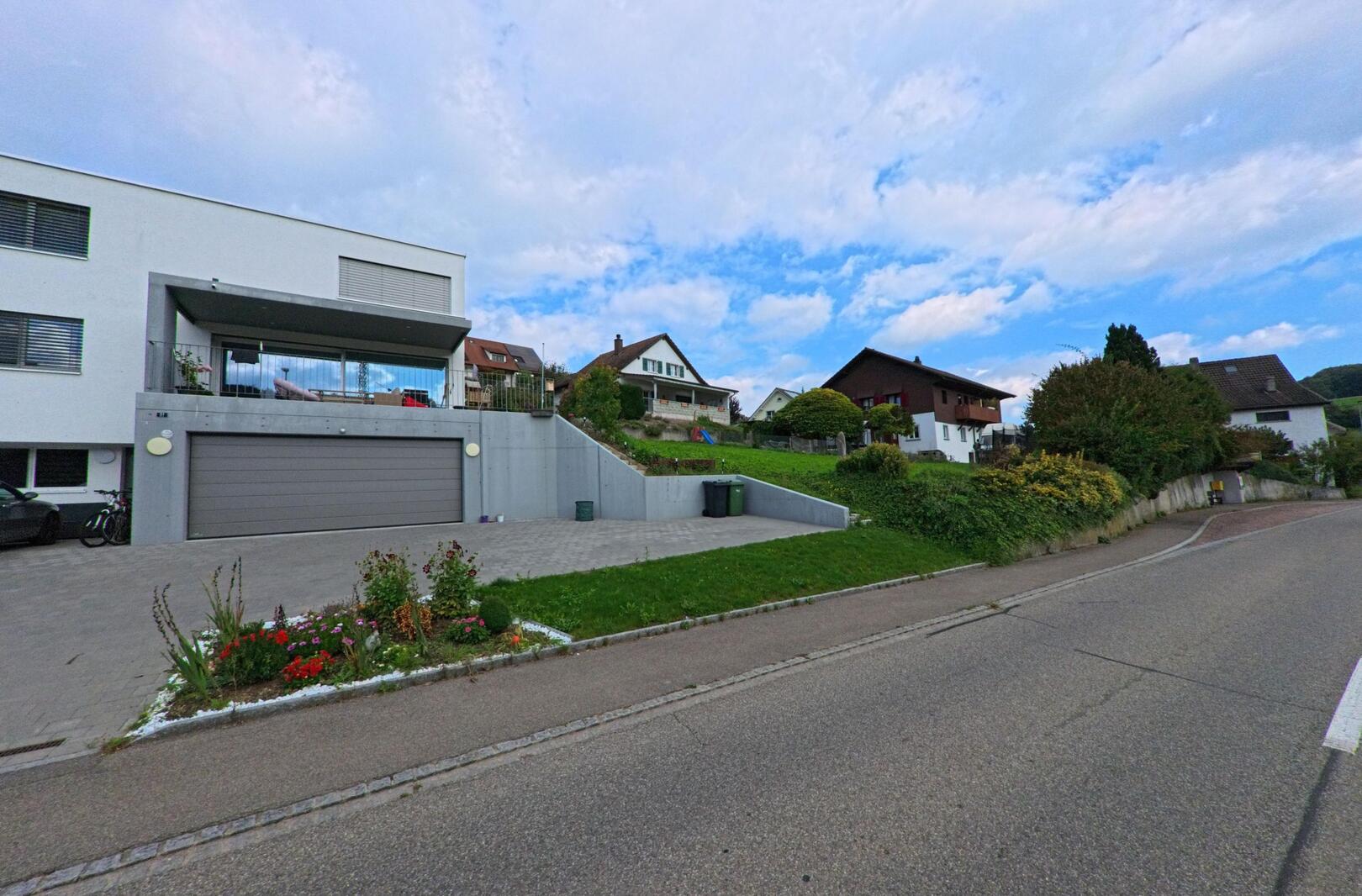 In Zuzgen (4315, Kanton Aargau) stehen moderne und traditionelle Häuser an einer leicht ansteigenden Strasse mit gepflegten Gaerten und weitem Himmel.