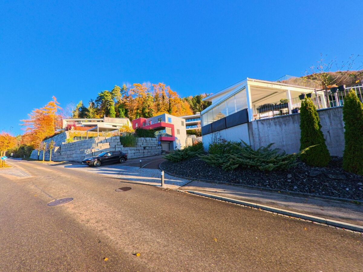 Am Hang gelegene moderne Wohnhaeuser praegen eine ruhige Quartierstrasse in Zuzwil (9524, Kanton St. Gallen) mit Blick auf herbstlich gefaerbte Baeume.