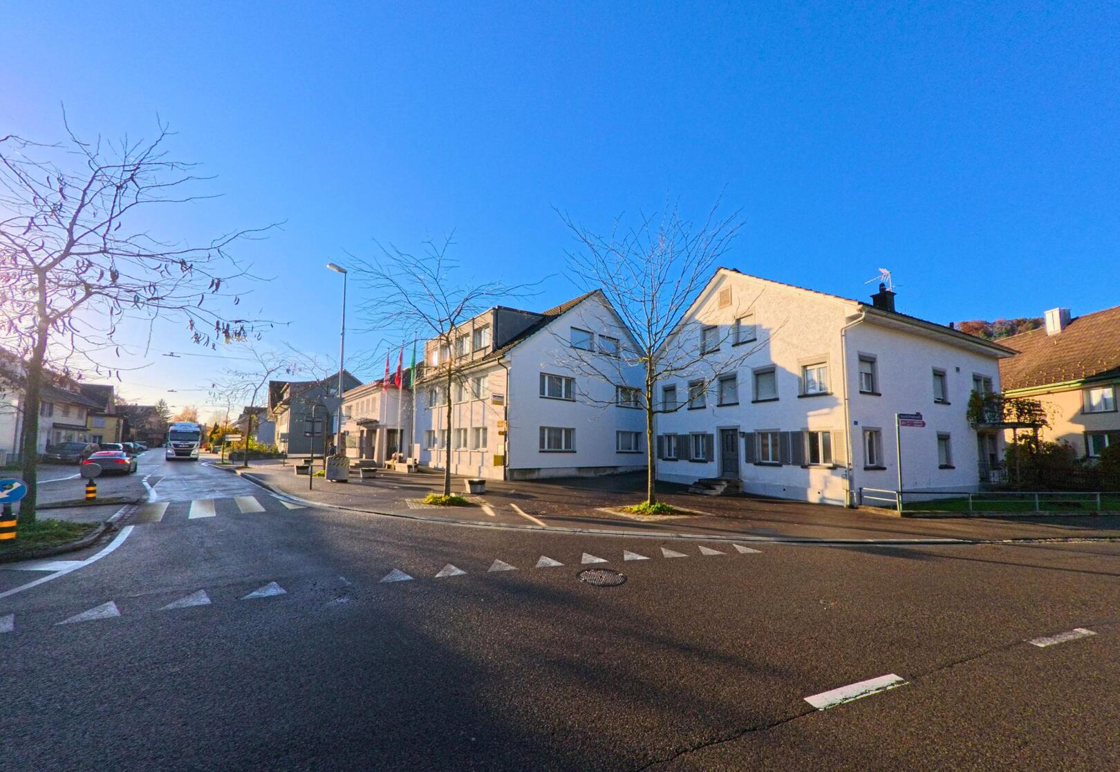 An einer ruhigen Strassenkreuzung in Zuzwil (9524, Kanton St. Gallen) stehen helle Wohnhaeuser mit kahlen Baeumen unter blauem Himmel am Morgenlicht.