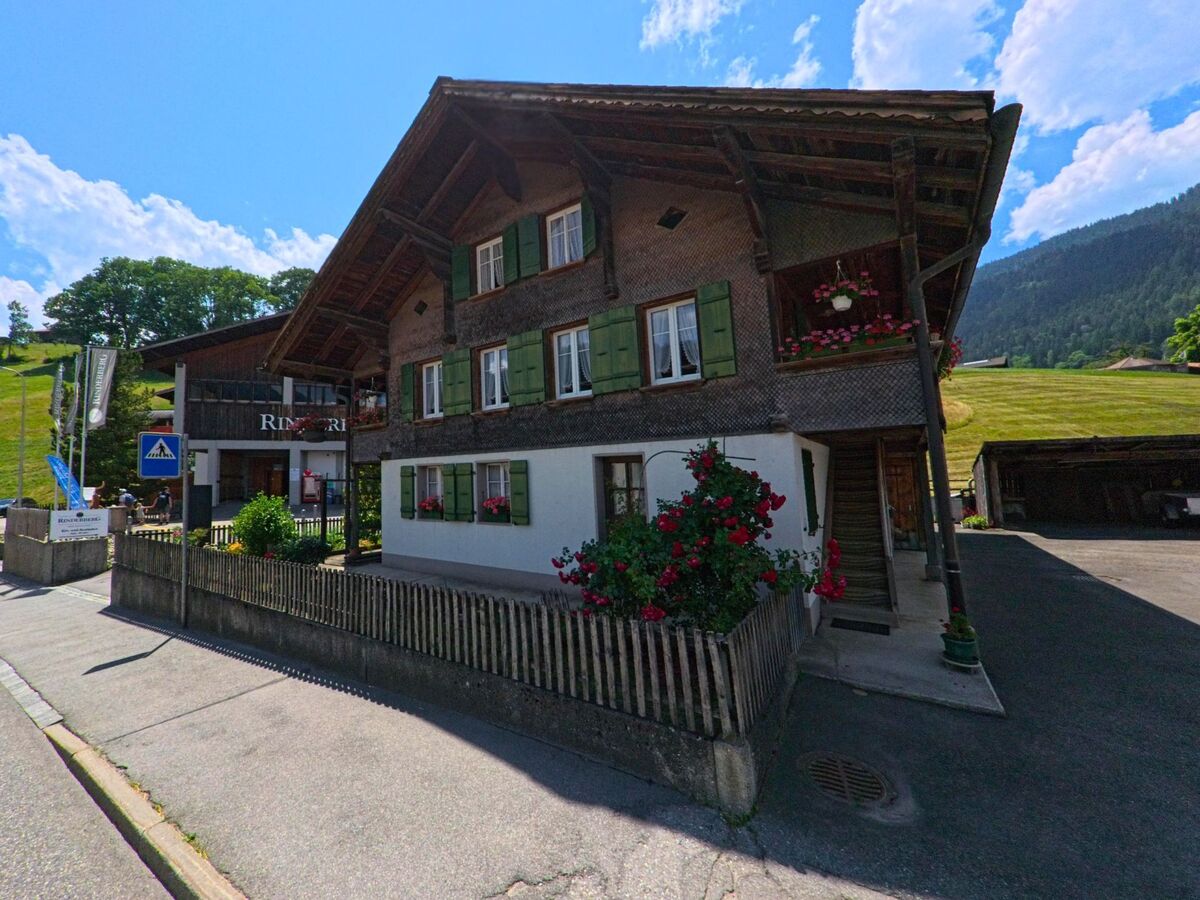 In Zweisimmen (3770, Kanton Bern) steht ein traditionelles Wohnhaus mit gruenen Fensterlaeden und Blumenschmuck direkt an einer Strasse mit Fussgaengerstreifen, dahinter liegt ein sanft ansteigender Hang.