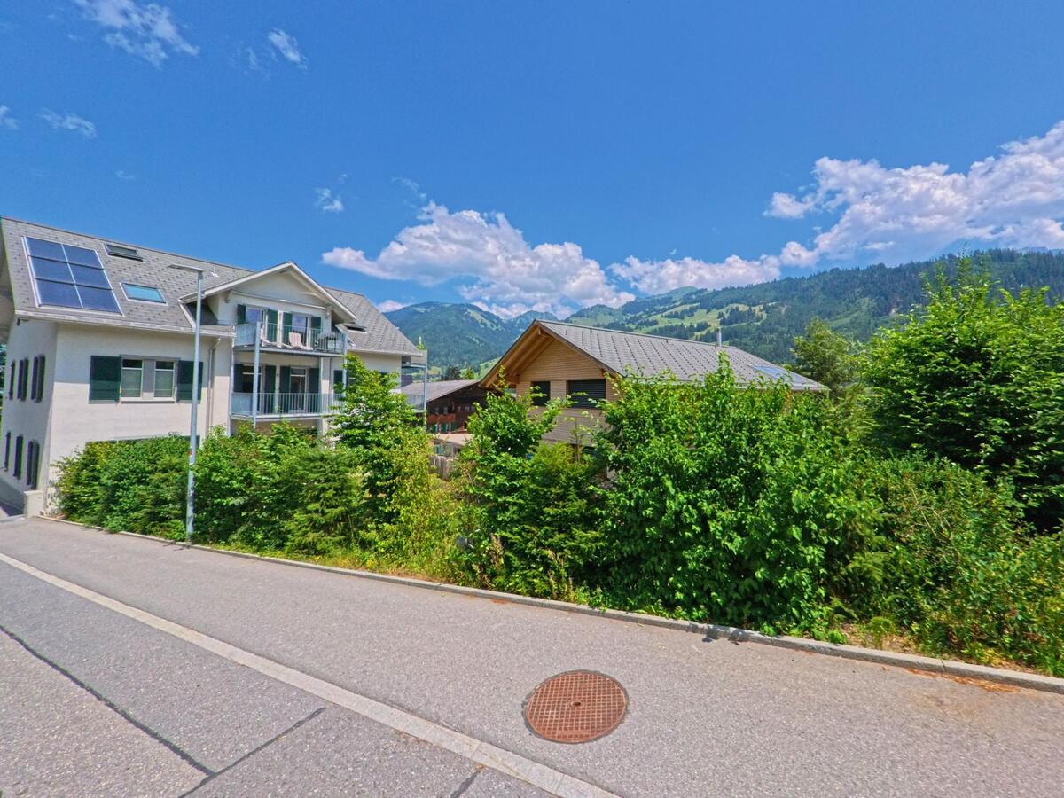 Zwei Wohnhaeuser mit Blick auf bewaldete Berghaenge stehen in Zweisimmen (3770, Kanton Bern) an einer ruhigen Quartierstrasse, umgeben von dichter Vegetation und blauem Himmel.