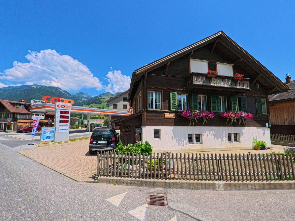 In Zweisimmen (3770, Kanton Bern) steht ein traditionelles Holzhaus mit blühenden Balkonkaesten direkt neben einer Coop-Tankstelle mit Blick auf die nahen Alpenhaenge.
