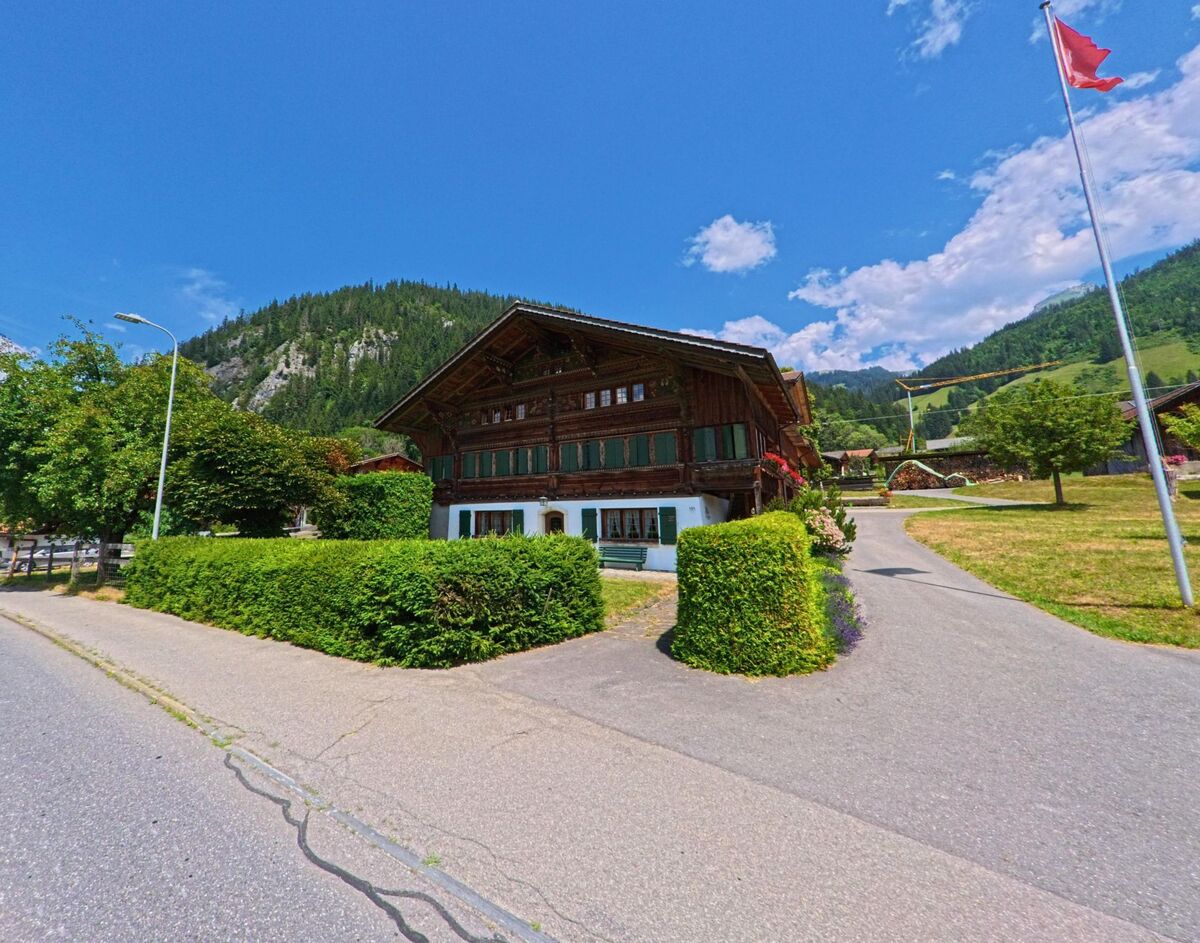In Zweisimmen (3771, Kanton Bern) steht ein gepflegtes Holzhaus mit grünen Fensterläden am Rand einer Wohnstrasse, umgeben von Hecken, Bäumen und einer Fahne im Vorgarten.