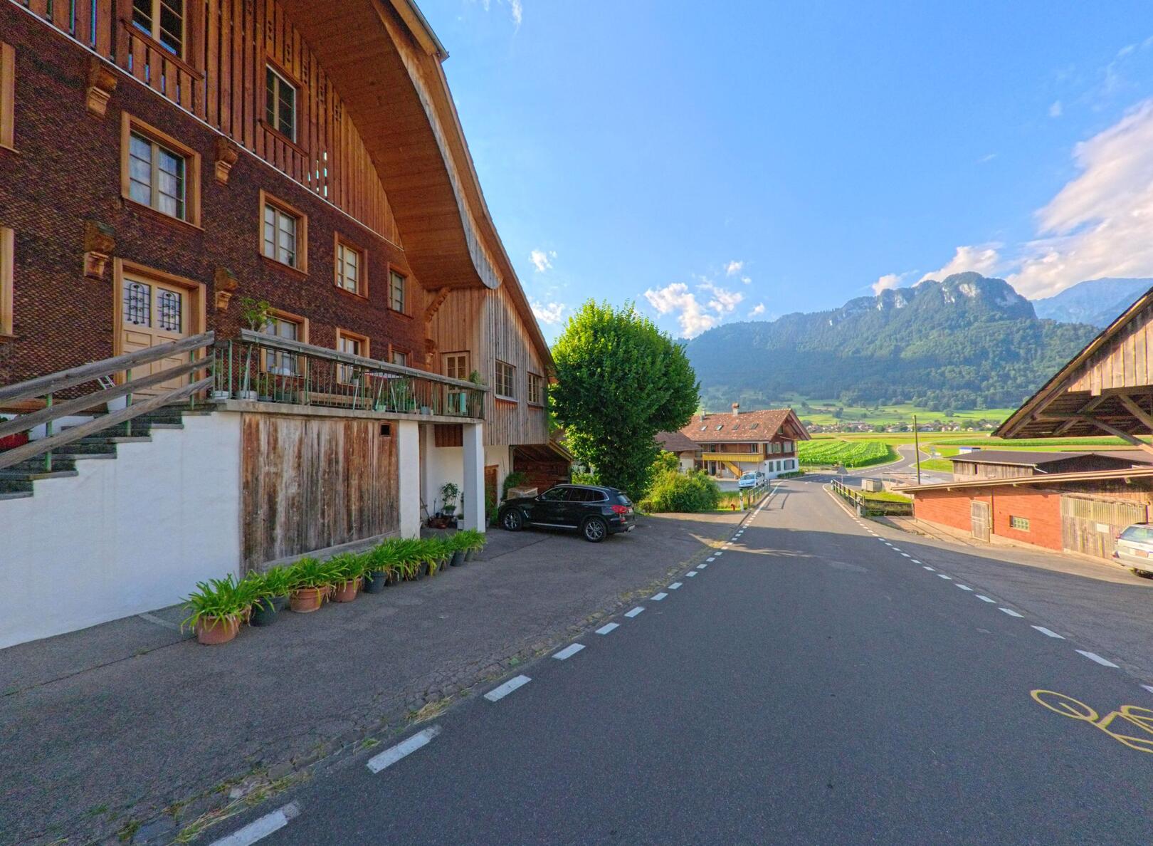 In Zwieselberg (PLZ 3645, Kanton Bern) fuehrt eine asphaltierte Strasse mit Fahrradmarkierung an traditionellen Bauernhaeusern vorbei in Richtung eines weiten Tals mit bewaldeten Huegeln.