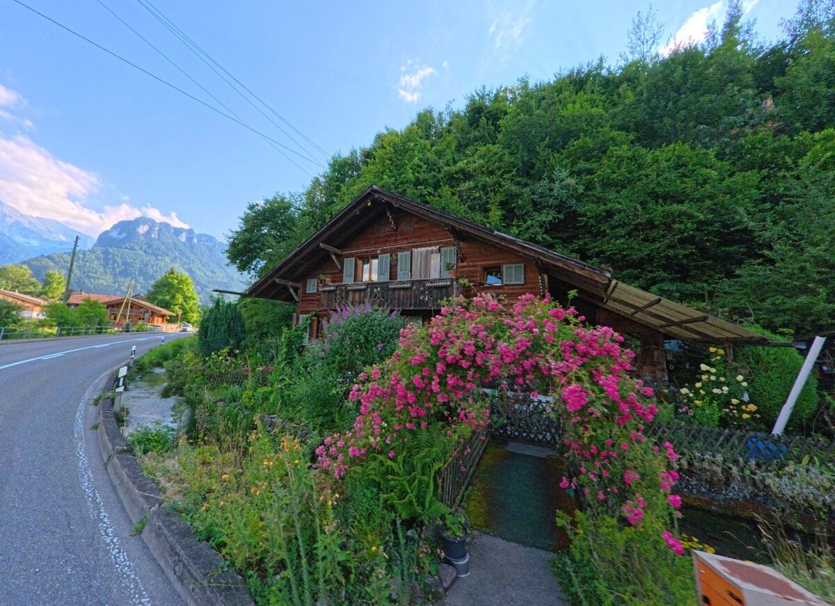 In Zwieselberg (PLZ 3647, Kanton Bern) steht ein traditionelles Holzhaus mit bunt blühendem Vorgarten direkt an einer kurvigen Strasse vor bewaldetem Hang.